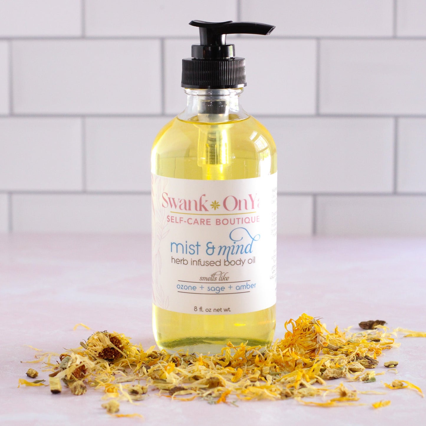 Bottle of 'Mist & Mind' body oil with a pump on a white surface with dried flowers.