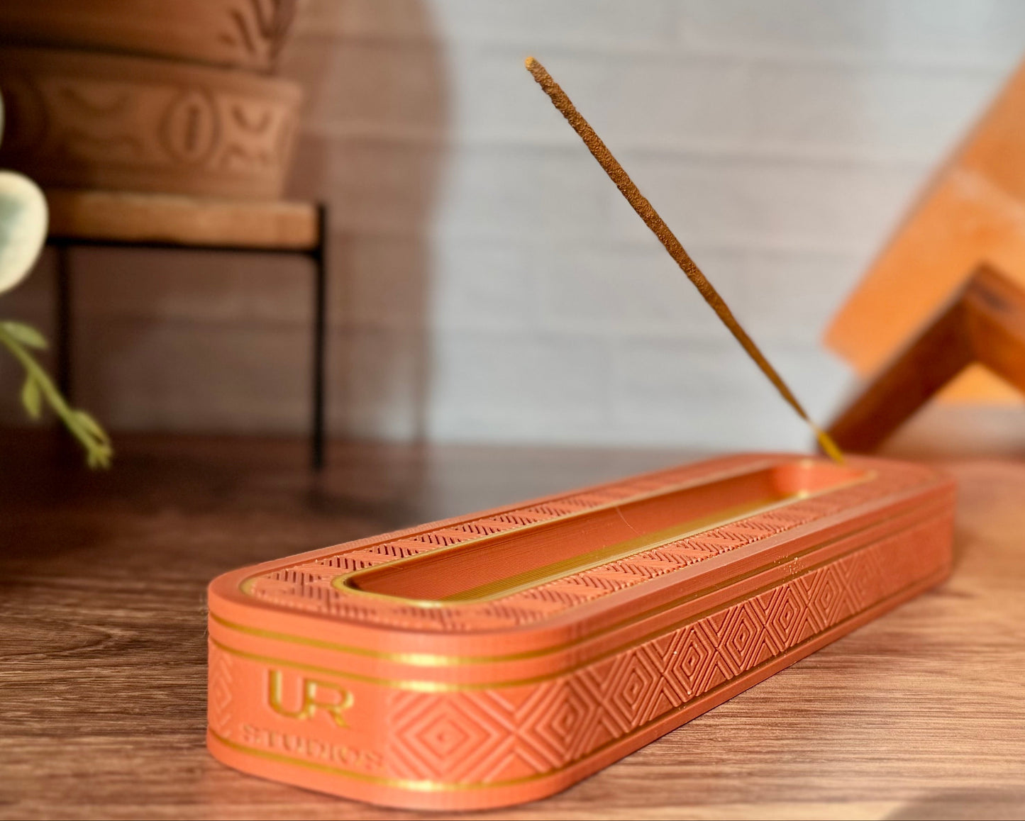 Incense stick holder with incense sticks on a wooden surface, with a plant and decorative items in the background.