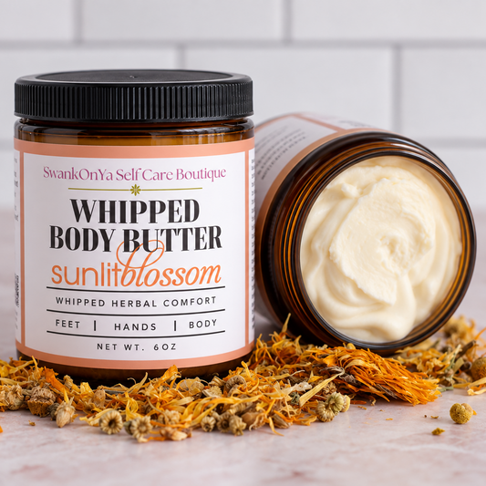 A jar of SwankiOnYa Self Care Boutique Whipped Body Butter labeled 'sunlitblossom' stands upright next to an open jar showing the whipped cream inside, styled with dried orange flowers on a marble surface against a white subway tile background.